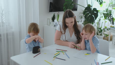Mom-and-her-two-sons-sitting-at-the-kitchen-table-drawing-colored-pencils-family-drawing-on-the-lawn-in-the-summer.-Boys-learn-to-draw,-and-mom-helps-with-drawing